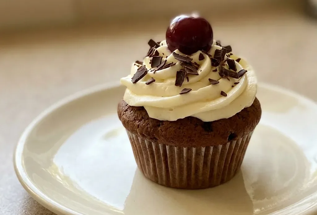 Overhead shot of a dark chocolate muffin topped with cream cheese frosting, dark chocolate shavings, and a sour cherry.