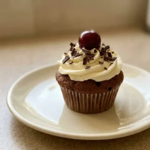 Overhead shot of a dark chocolate muffin topped with cream cheese frosting, dark chocolate shavings, and a sour cherry.