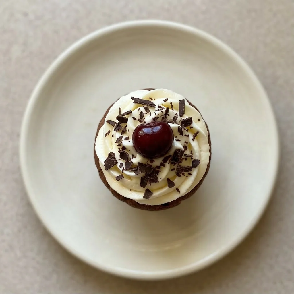 Plated muffin with a generous dollop of cream cheese frosting, scattered dark chocolate shavings, and a single sour cherry.