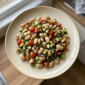 Looking down at a bowl of butter bean salad with cherry tomatoes, cucumber, red onion, parsley, and lemon wedges.