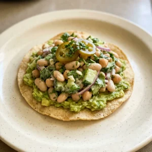 Looking down at a bowl of bean salad with cannellini beans, chickpeas, cilantro, lime wedges, jalapeño slices, and avocado chunks, with tostadas on the side.