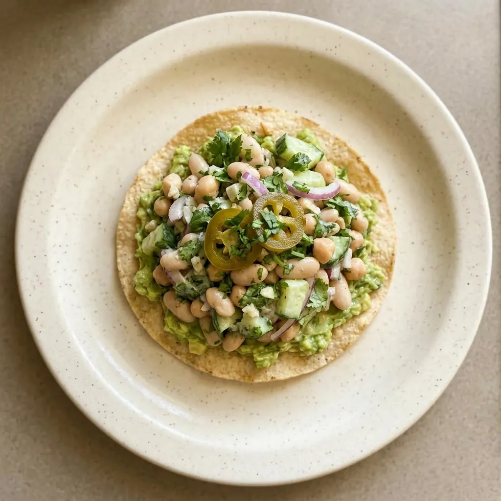 A plate of marinated cilantro lime bean salad topped with avocado chunks, jalapeño slices, and cilantro leaves, with tostadas around the edge.