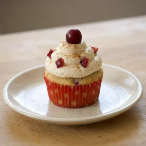 Bird's-eye view of a white chocolate-drizzled cupcake topped with a sugared cranberry and a cinnamon stick, with nutmeg specks visible.