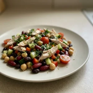 Top-down look at a mix of beans, chicken chunks, cherry tomatoes, cucumber slices, red bell pepper strips, and fresh herbs drizzled with olive oil and lemon juice.