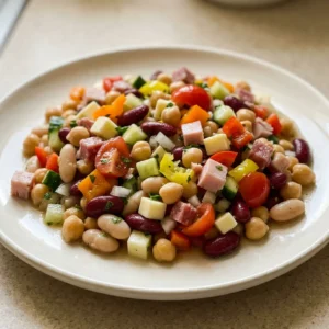 Top-down look at a dense bean salad with chickpeas, cannellini, kidney beans, cucumber, sweet onion, cherry tomatoes, bell peppers, banana peppers, provolone, turkey, and salami.