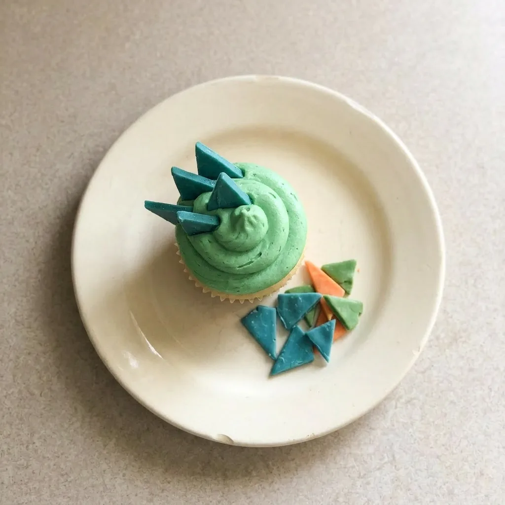 A serving of a dinosaur cupcake with green frosting, candy eyes, and sprinkles, viewed from the side.