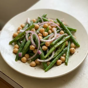 Looking down at a bed of green beans and chickpeas, scattered with red onion rings and parsley, drizzled with olive oil.