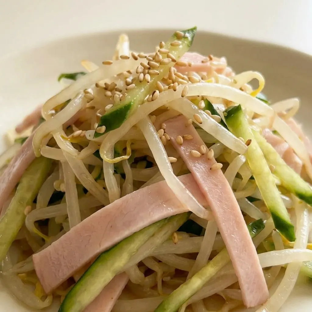Zoomed in on a mix of bean sprouts, cucumber, and ham, glistening with rice vinegar and soy sauce, sprinkled with sesame seeds.