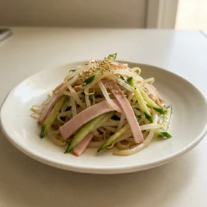 Bird's-eye view of a bed of bean sprouts topped with cucumber slices, ham strips, sesame seeds, and a drizzle of soy sauce and sesame oil.