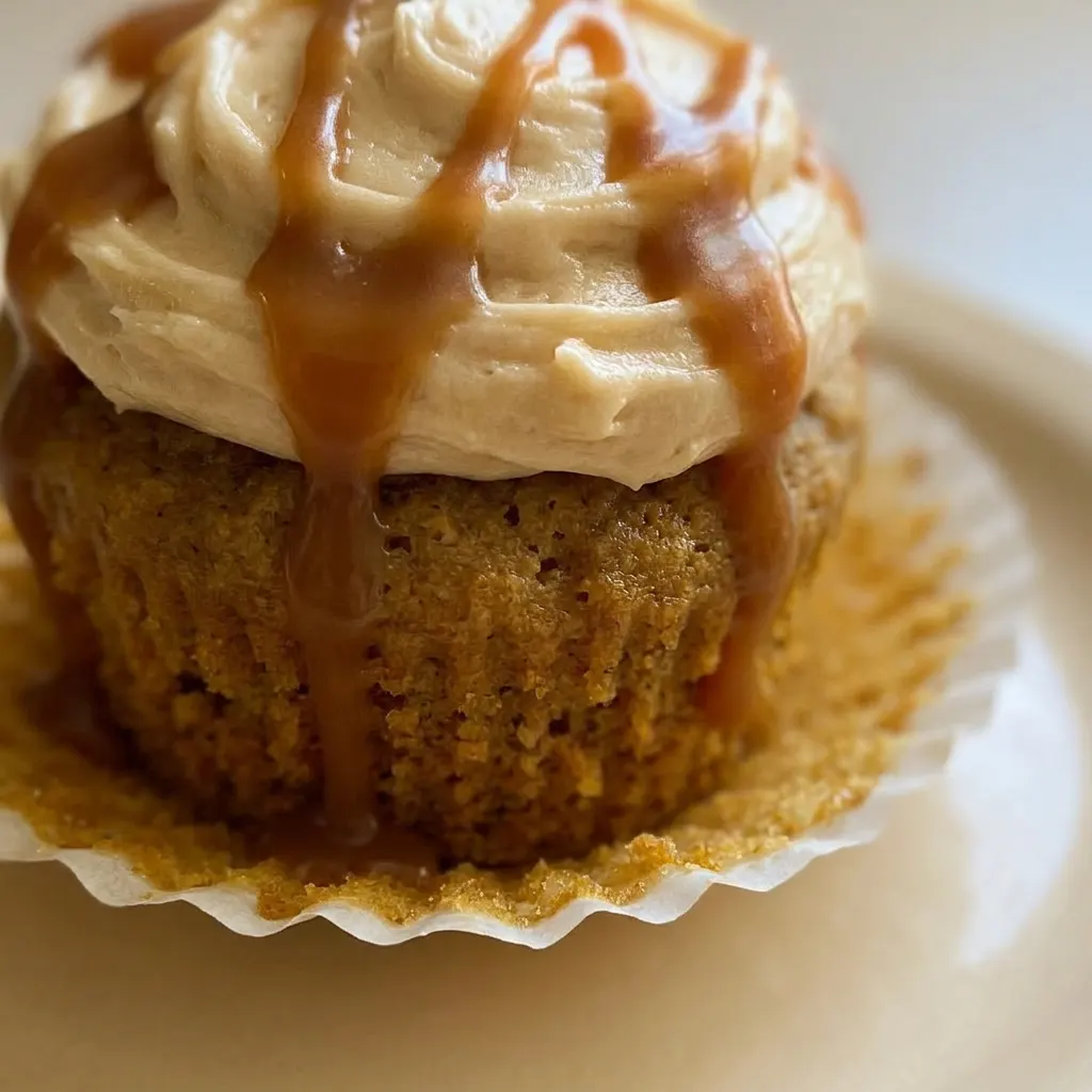 Close view of a pumpkin cupcake with caramel cream cheese frosting, topped with a caramel swirl and spice dusting.