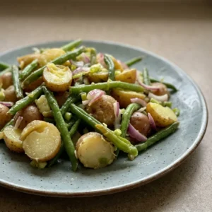 Looking down at a bowl of string bean and potato salad with red onion rings, lemon slices, and olive oil drizzle.