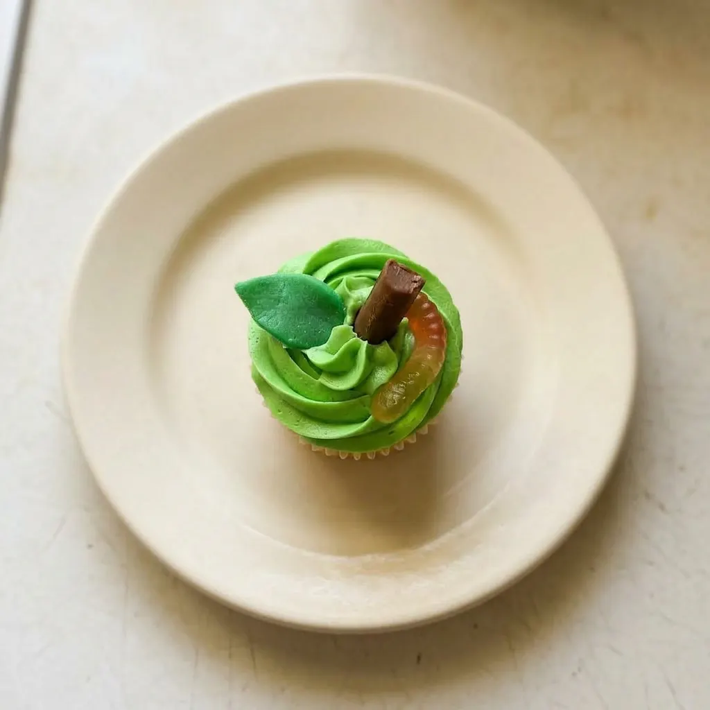 Plated apple cupcake with frosting, Tootsie Roll, Airhead strip, and gummy worms on top.
