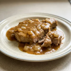 Looking down at a pork chop topped with sliced onions and a glossy brown gravy, with a pool of gravy on the side.