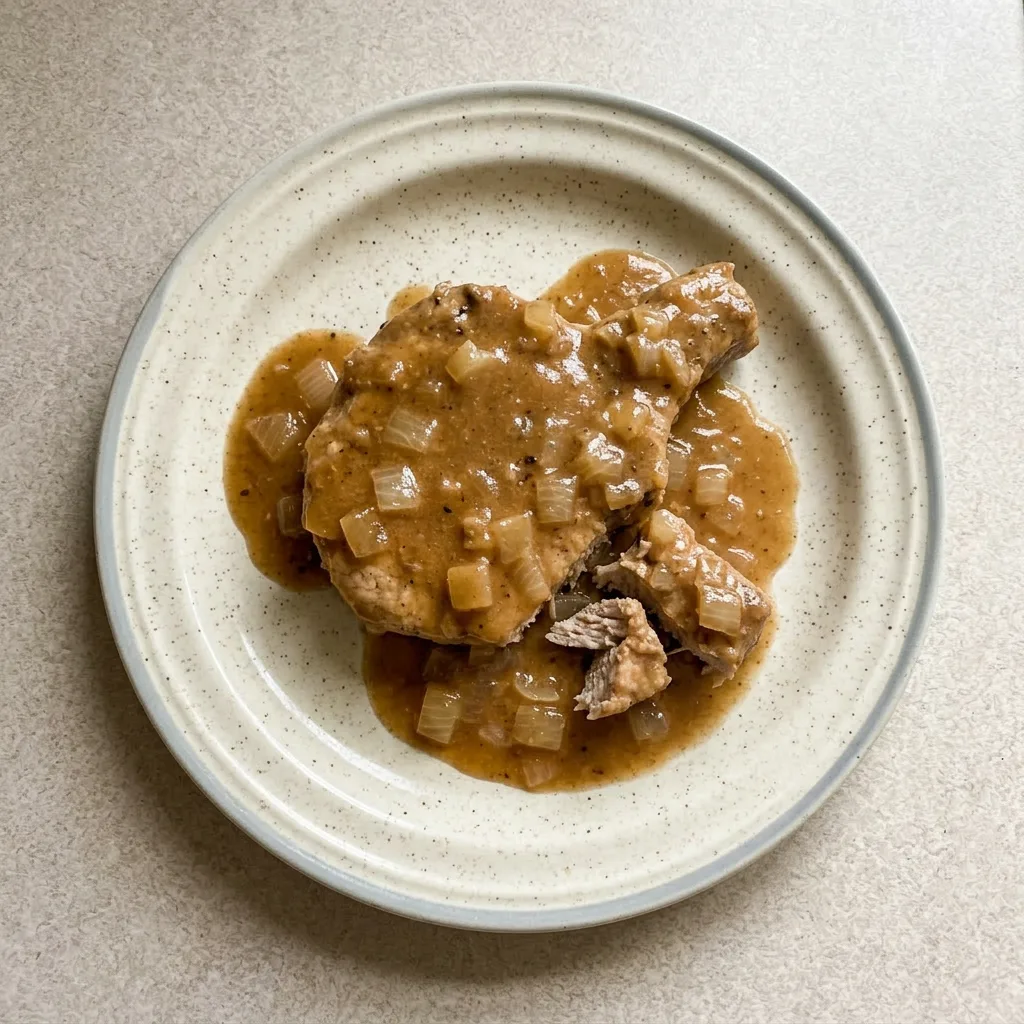 A plate of a single pork chop covered in rich gravy and soft onion slices, with extra gravy pooling around the meat.