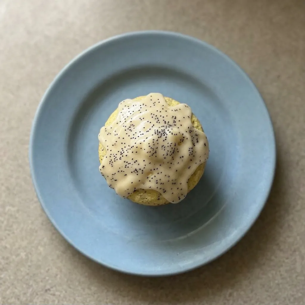 Plated muffin with a lemon glaze drizzle and poppy seeds visible on the top, golden-brown edges, and a soft crumb.