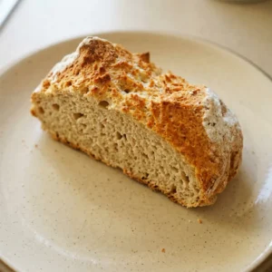 Overhead shot of a round soda bread loaf with a cross cut on top, golden brown crust dotted with white and whole wheat flour specks.