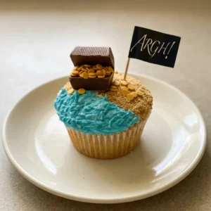 Looking down at a vanilla cupcake with blue frosting, gold sprinkles, and a Hershey's nugget on top.