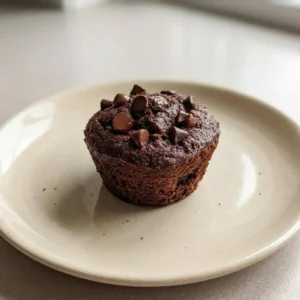 Overhead shot of a chocolate muffin with a domed top studded with chocolate chips on a dark surface.