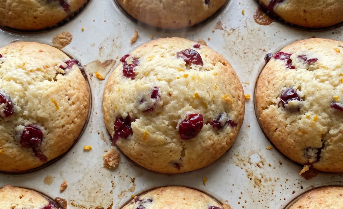 Cranberry orange muffins with golden tops, dried cranberries and orange zest visible, fresh orange slices alongside.