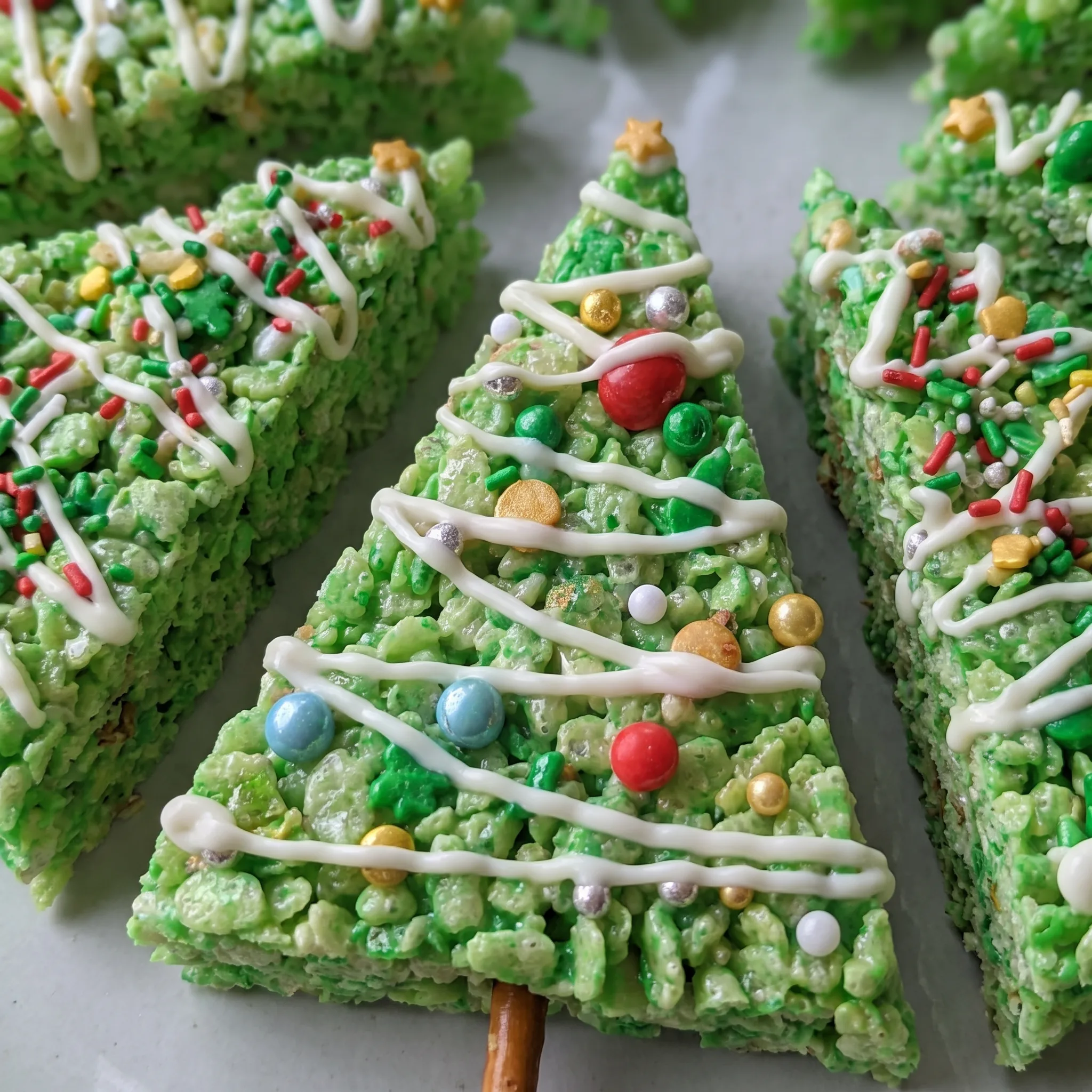 Crispy green Rice Krispie tree with a white chocolate drizzle and red, green, and white sprinkles, resting on a pretzel stick trunk.