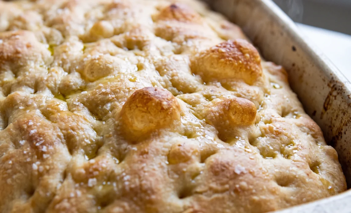 Sourdough focaccia with a golden crust, dimpled surface, glossy olive oil, and scattered flaky sea salt.