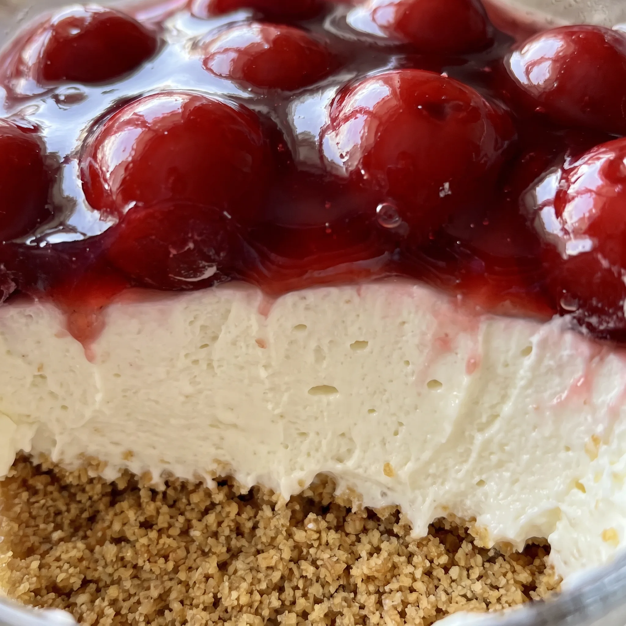 Glossy cherry pie filling with whole cherries and stems sits over smooth cream cheese and whipped cream layers, graham crumbs between.