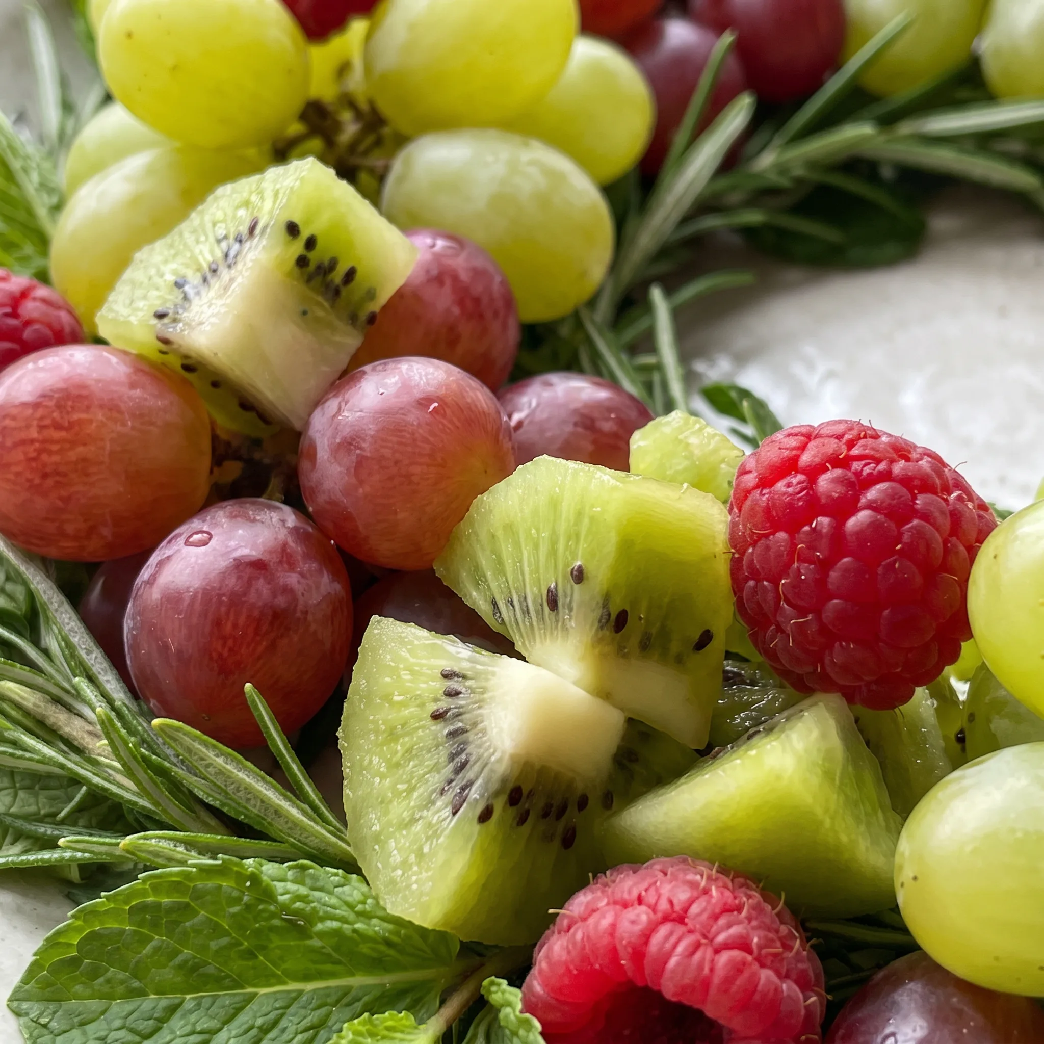 Glossy raspberries and halved green grapes tucked among kiwi slices and rosemary sprigs in a fruit wreath.