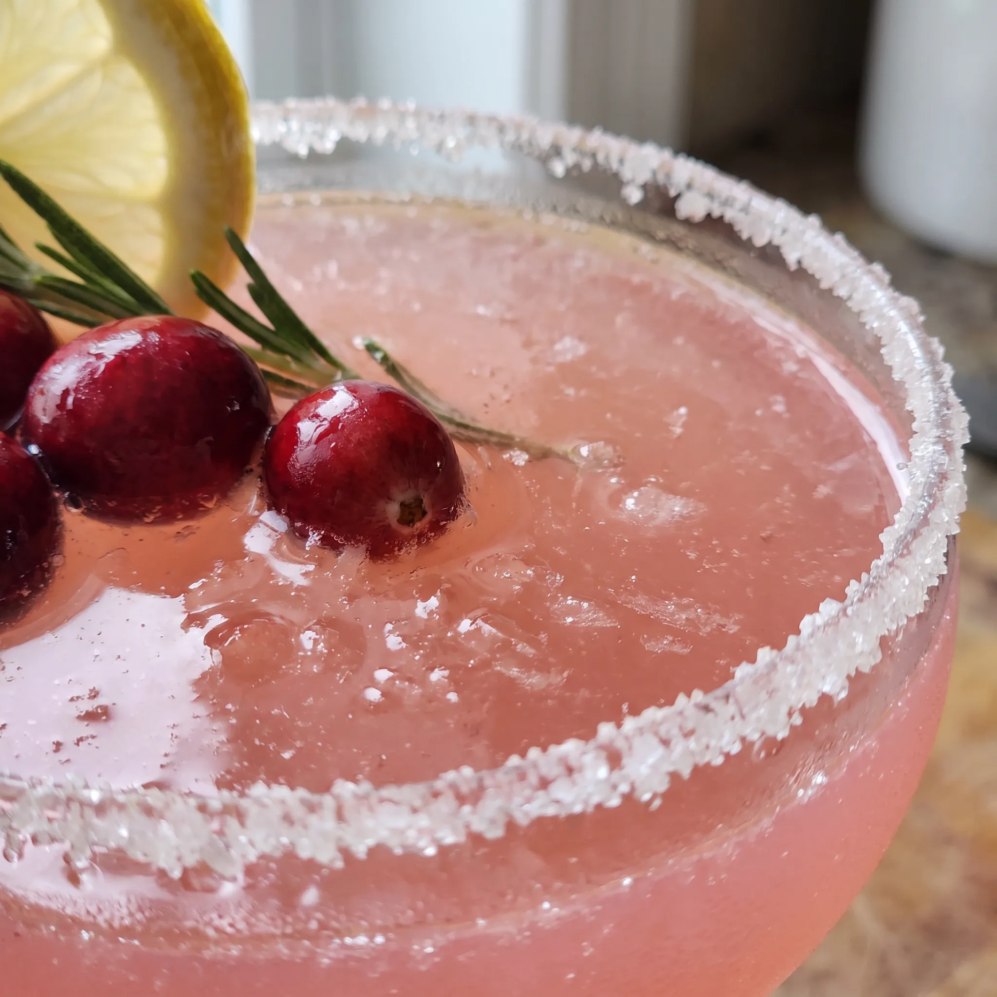 Glossy whole cranberries and a curled lemon peel beside a rosemary sprig in a pale pink cocktail.