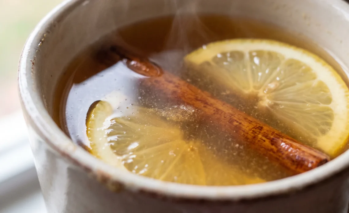 A hot toddy in a glass mug with amber tea and whiskey, a lemon slice floating on top and a drizzle of honey.