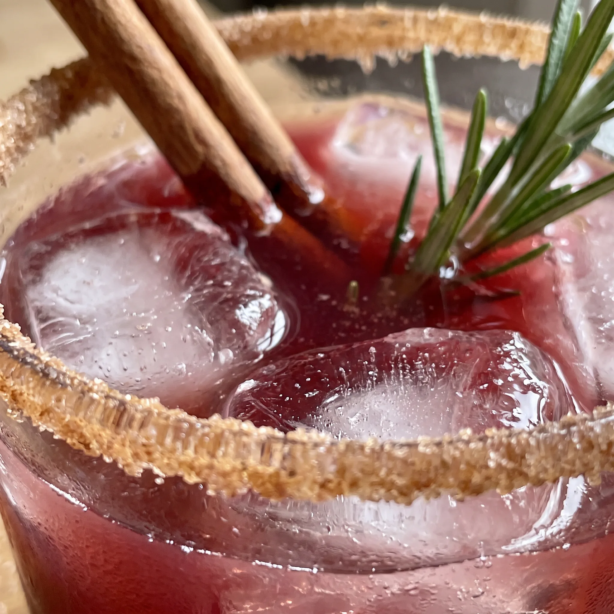 Red pomegranate margarita with a cinnamon-dusted rim, a cinnamon stick, and a fresh rosemary sprig, showing a smooth, chilled texture.