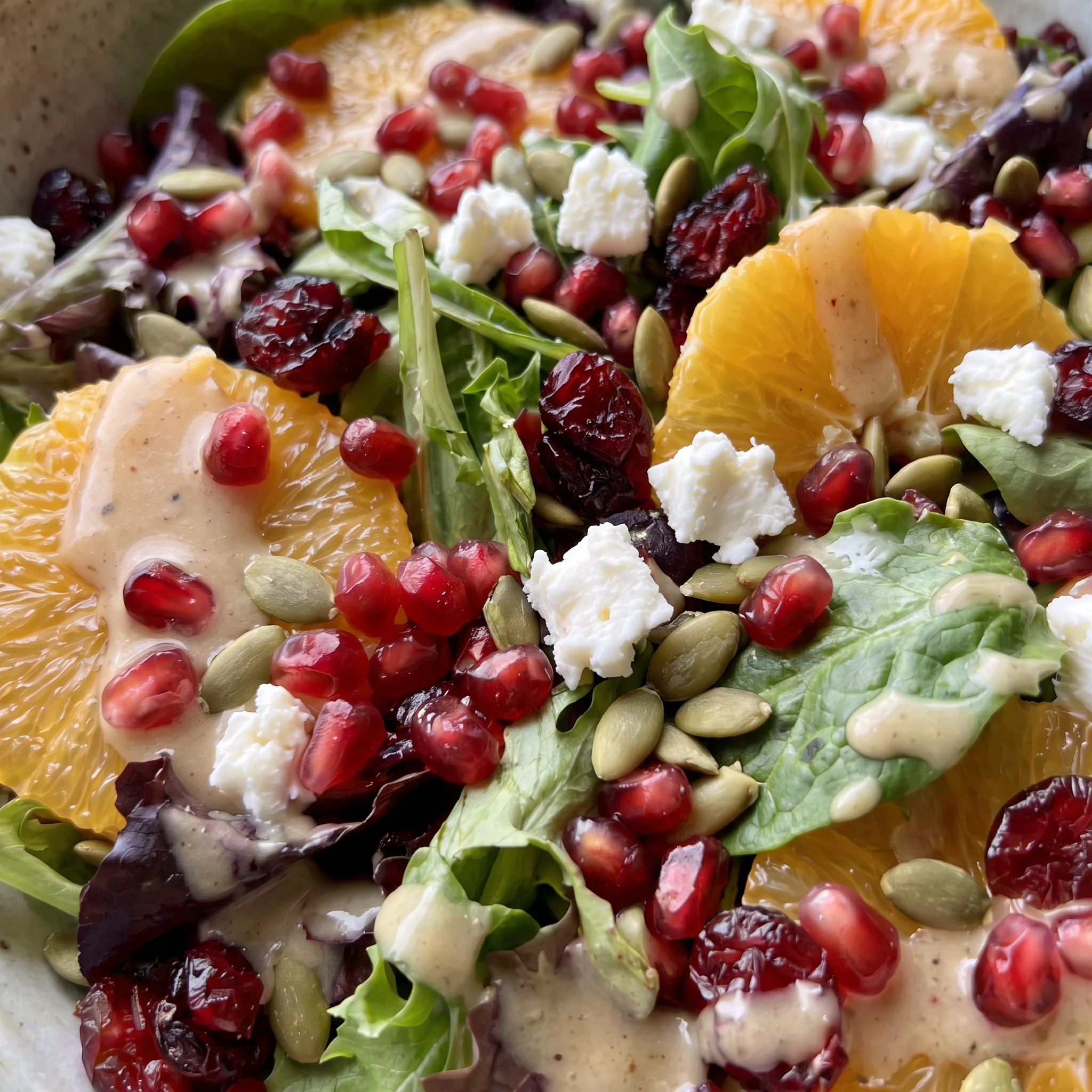 Glossy pomegranate seeds and pepitas scattered over feta crumbles and orange slices on dark leafy greens.